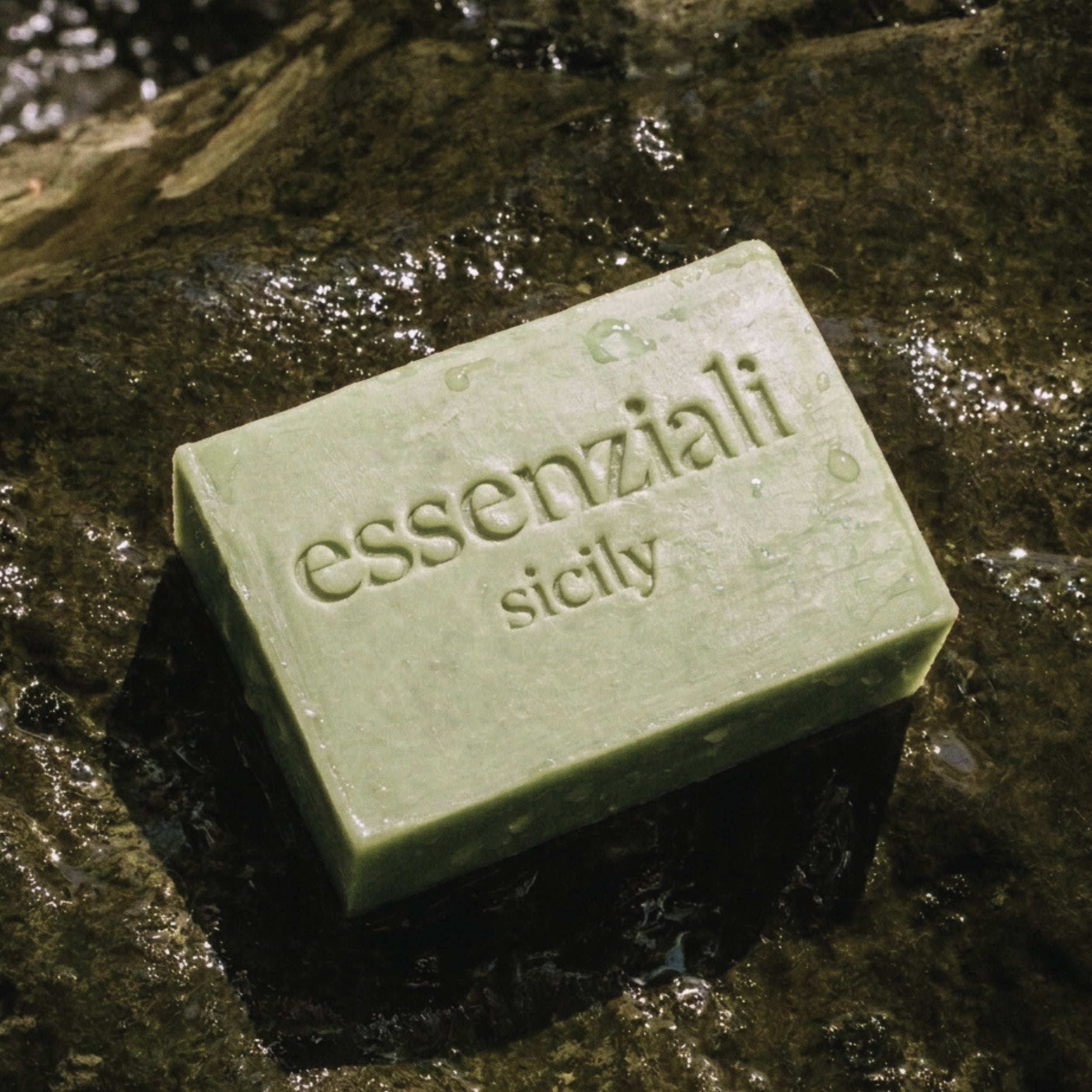 Green bar of soap with 'essenziali sicily' branding on a wet stone surface