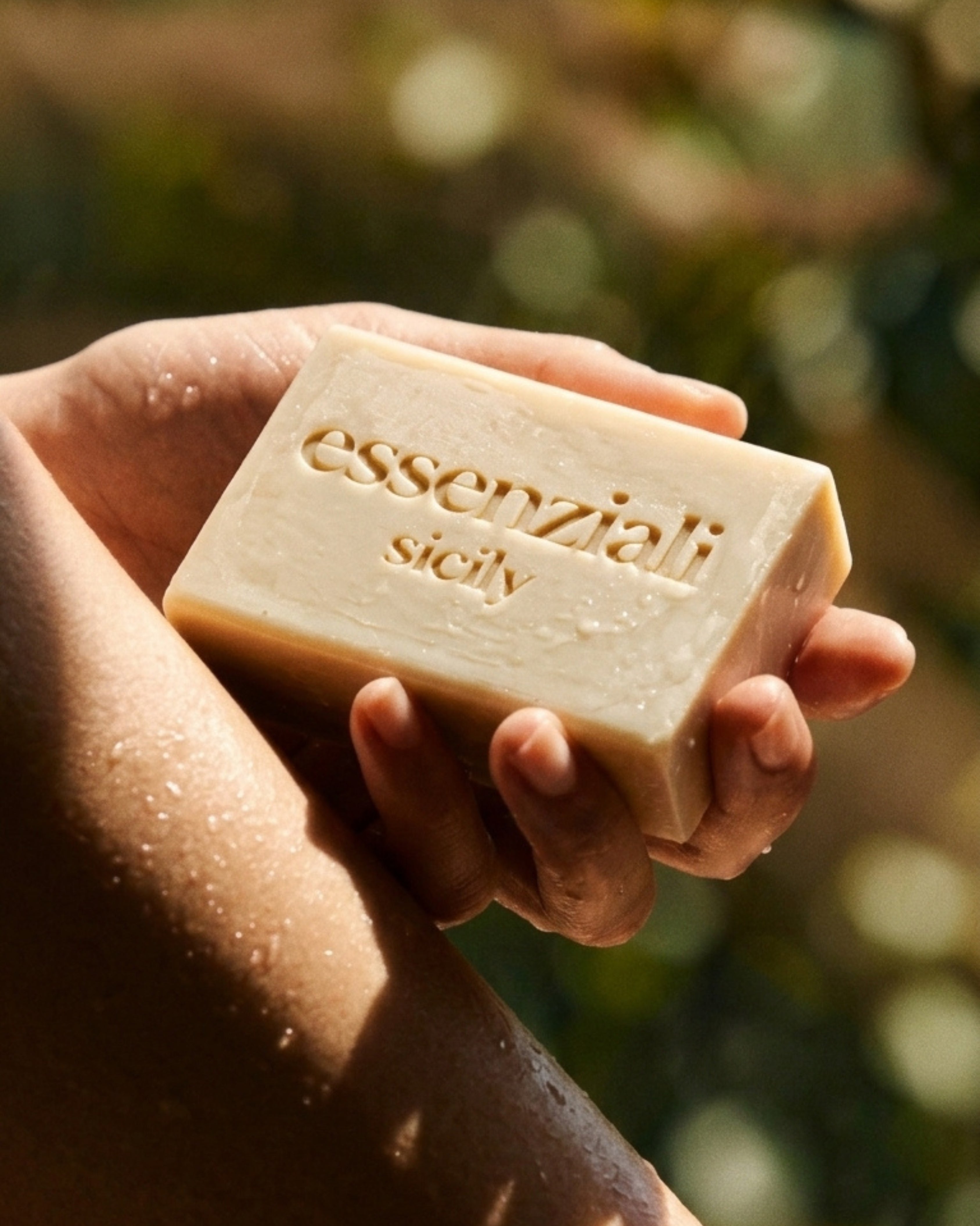 Hand holding a bar of soap with 'essenziali sicily' branding against a blurred natural background. Natural handmade soap from Italy. 