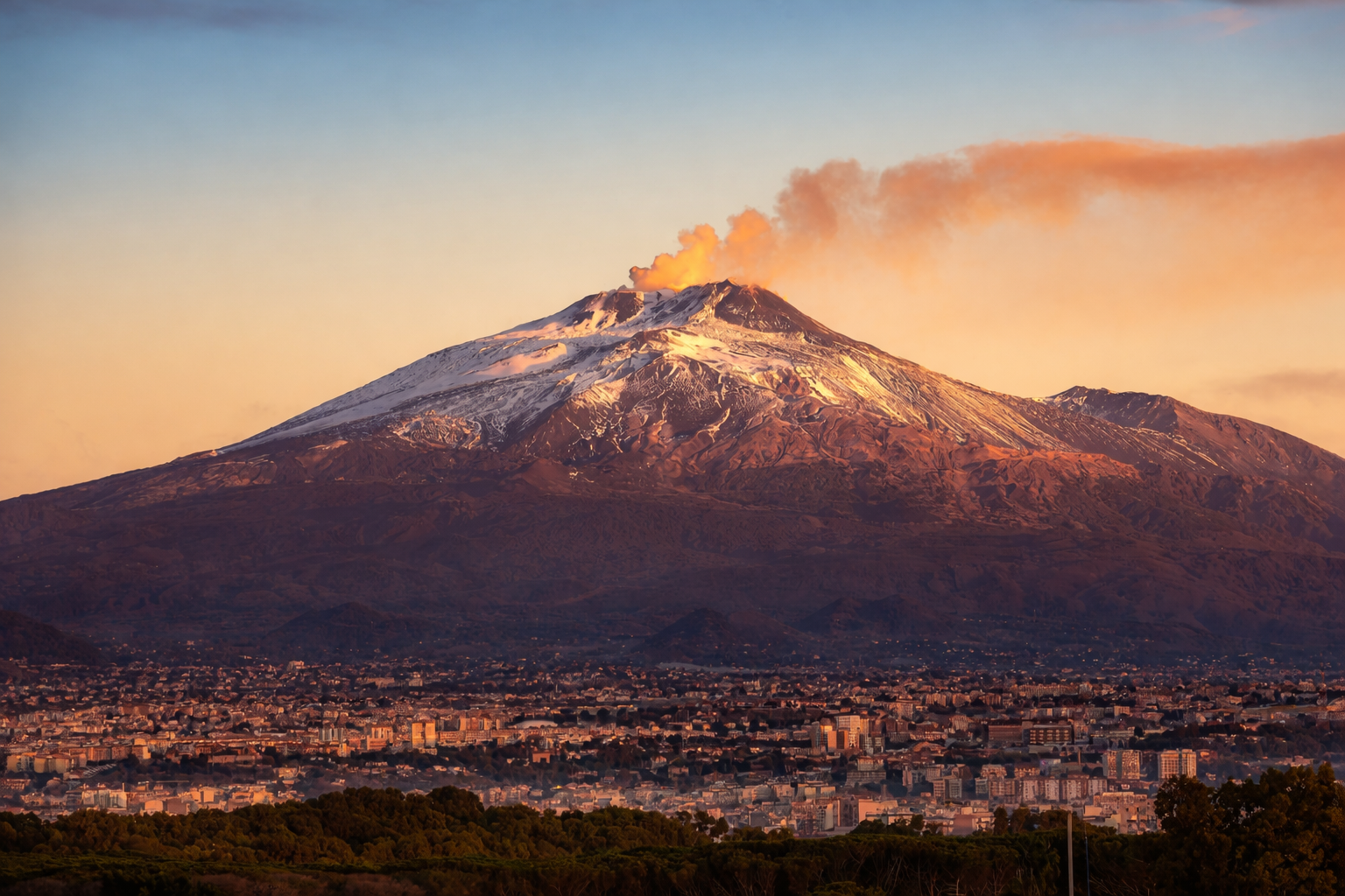 Mount Etna in Sicily with snow caps and cityscape below at sunset.