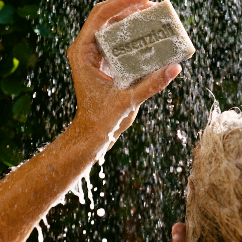 Hand holding a lathered Essenziali soap bar beneath falling water outdoors